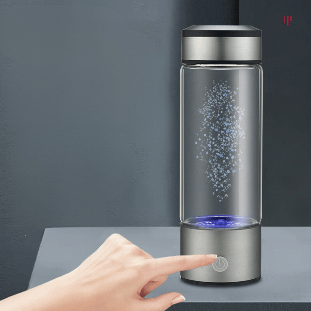 Glass water bottle with a silver base and black lid on a dark surface, with a hand pointing at the power button.